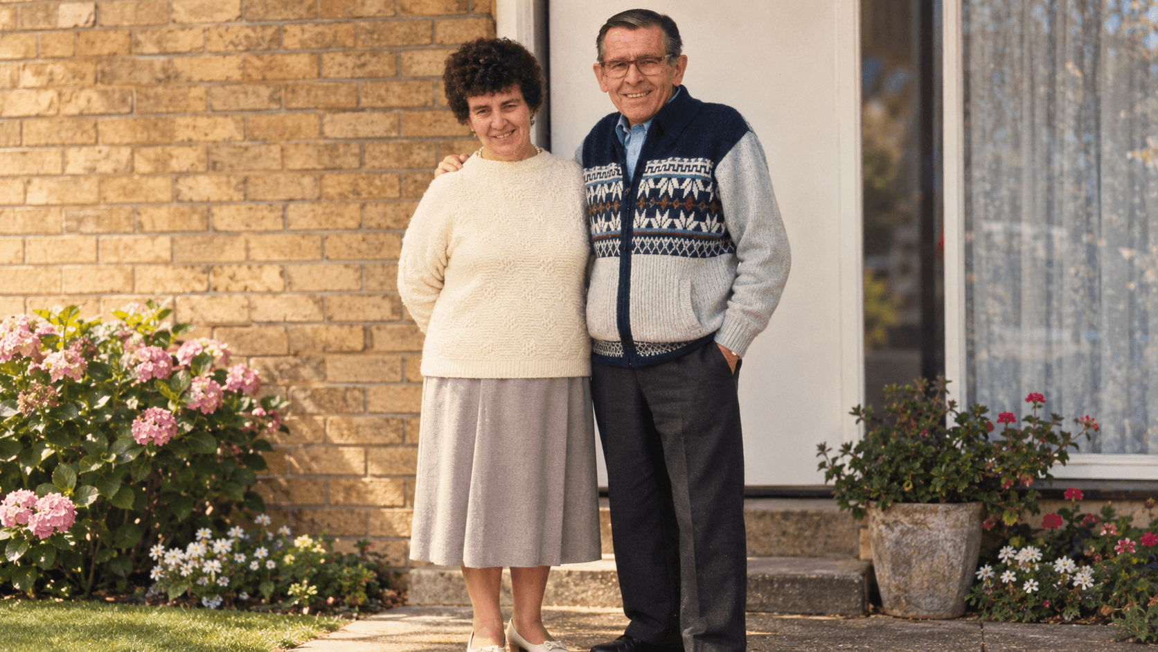 Sheila May Tasker standing with her husband Stanley outside their home, surrounded by flowers